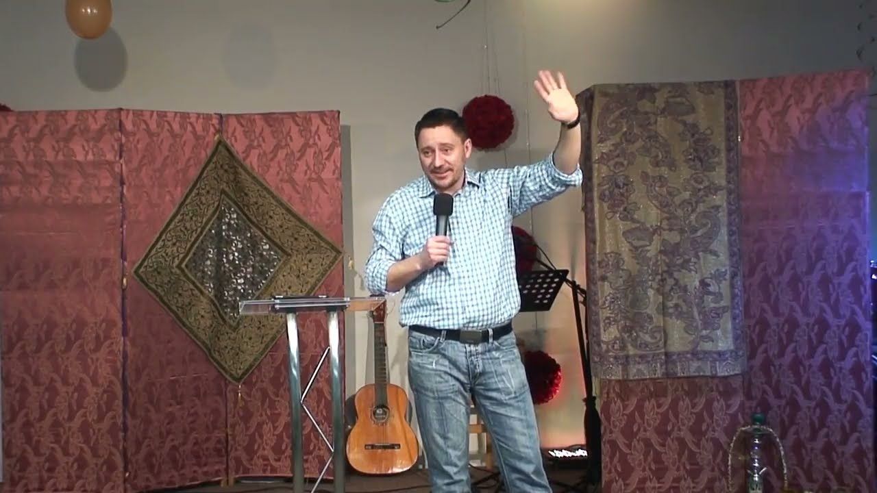 A man in a checked shirt raising an open hand and holding a microphone in front of fabric screens.