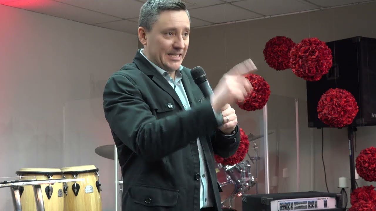 Ruslan Murin in a dark jacket preaching into a microphone in front of red flower decorations.