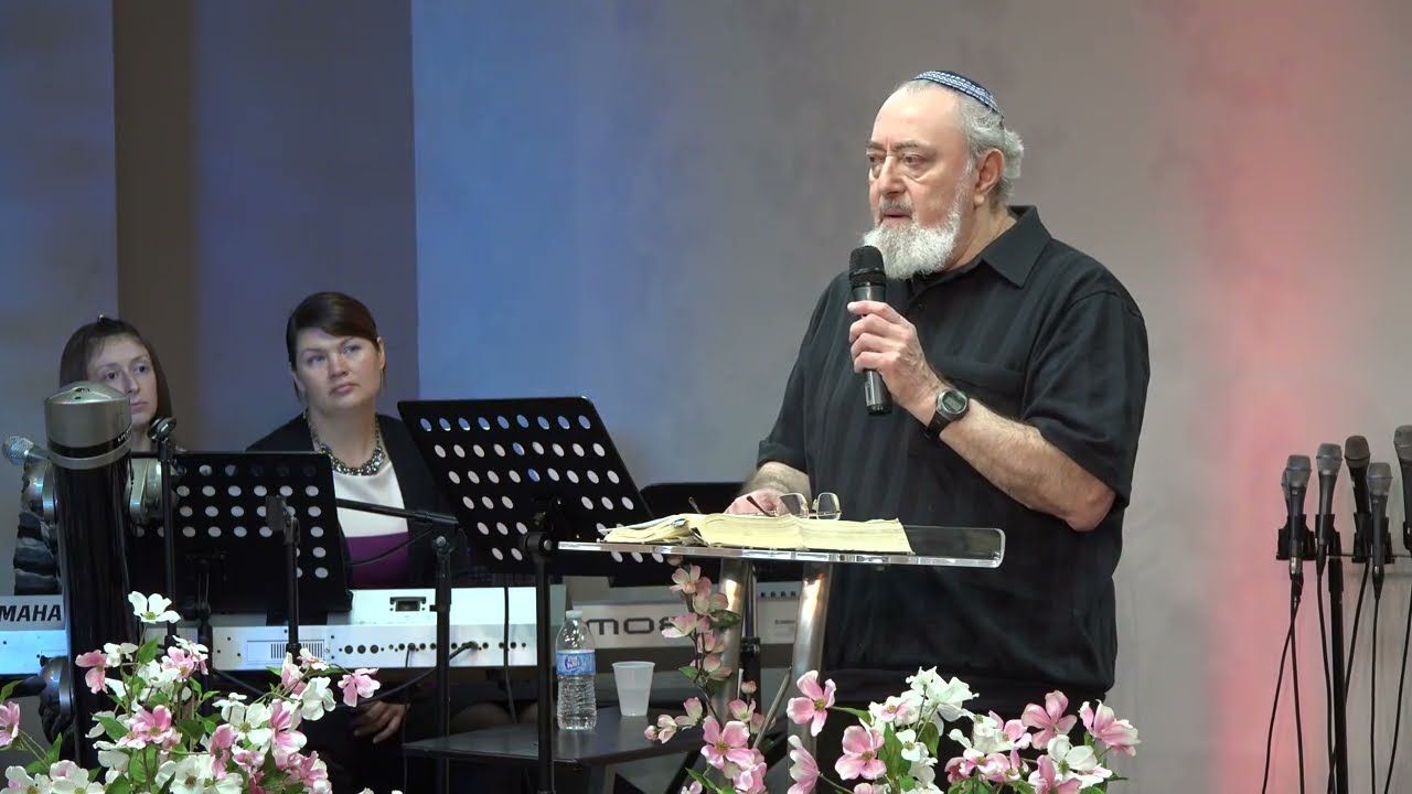 A man with a white beard and a kippah speaking into a microphone at a lectern, two women seated behind.