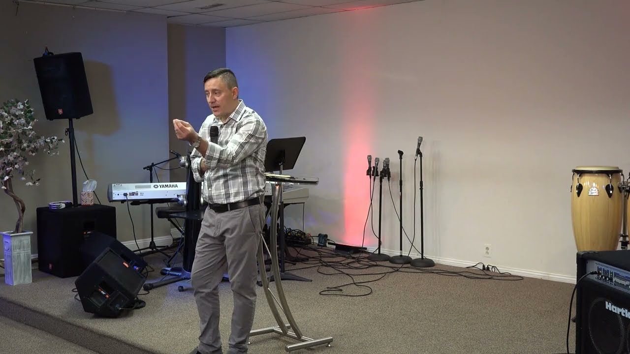 Ruslan Murin in a plaid shirt preaching with a microphone beside a lectern and gesturing with one hand.