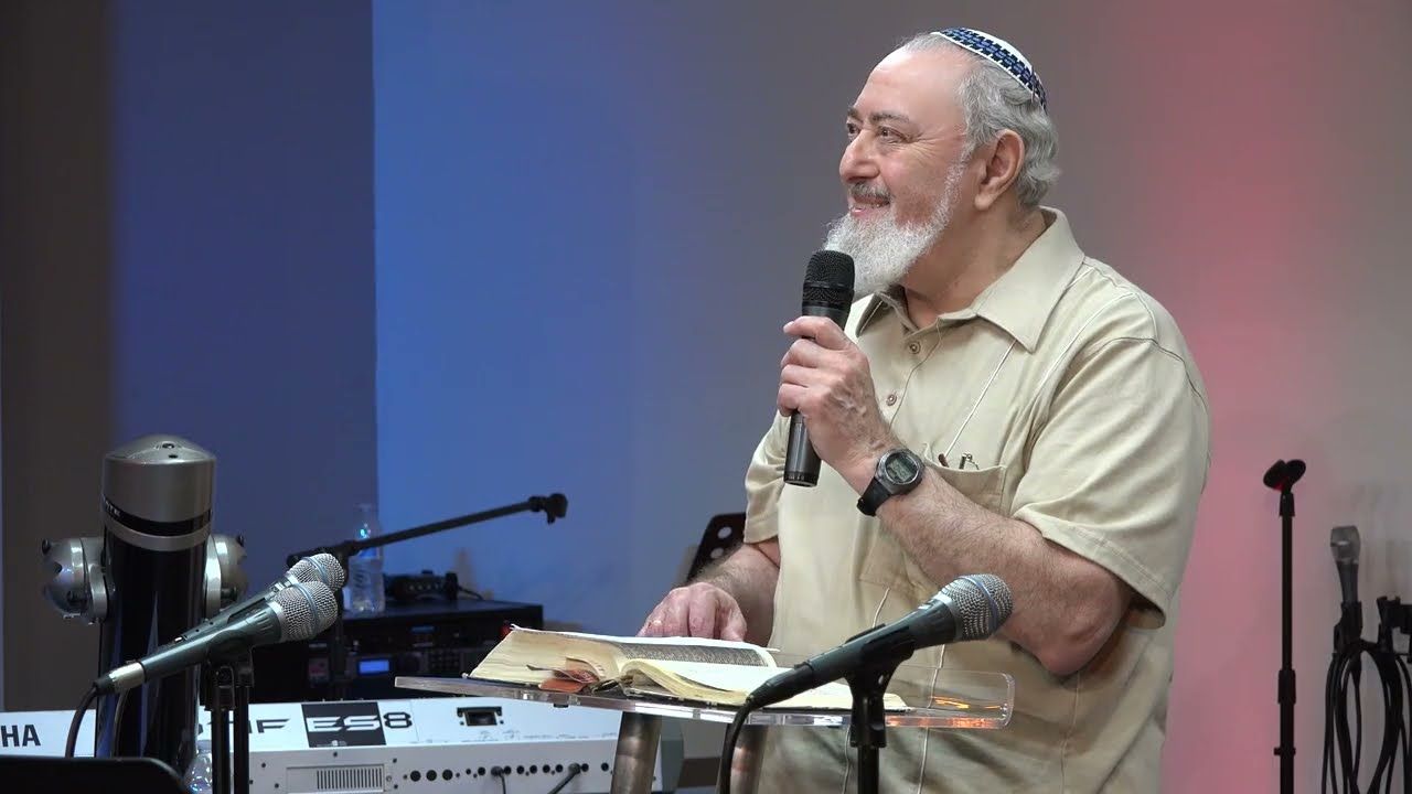 A man with a white beard and a kippah holding a microphone and smiling at a lectern with an open book.