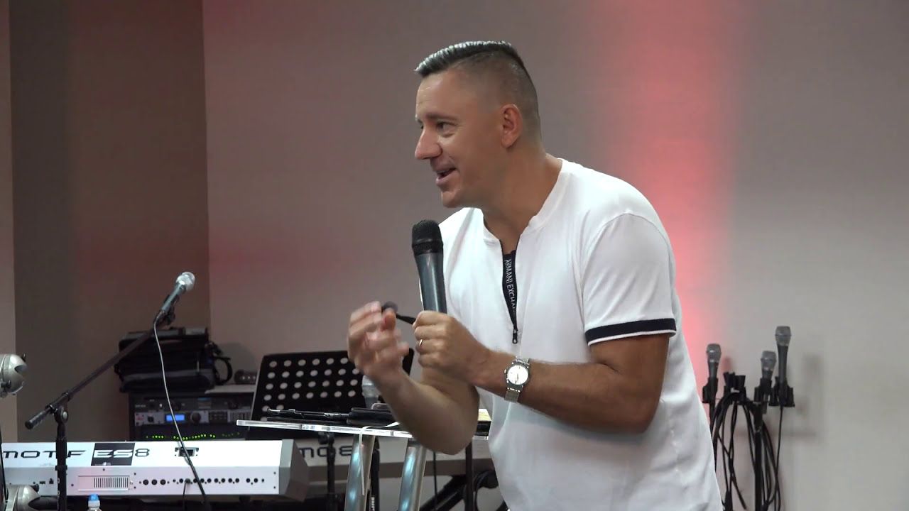 Ruslan Murin in a white T-shirt preaching with a microphone and gesturing with one hand.