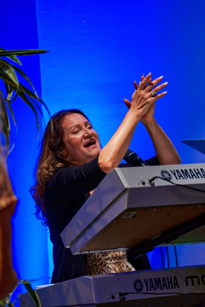 A woman at a keyboard singing with both hands raised