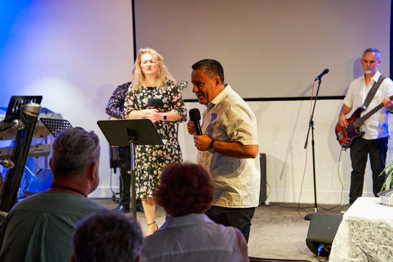 A man with a microphone and a woman on the stage, a bass player behind them, seen over the congregation